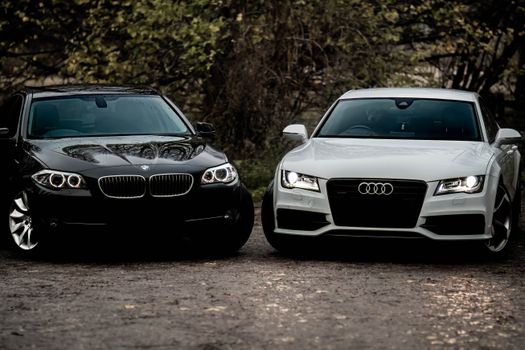 Black BMW and white Audi luxury sedans parked outdoors on a gravel path.