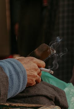 Hand holding a cigarette and smartphone, surrounded by smoke, in an intimate close-up shot.