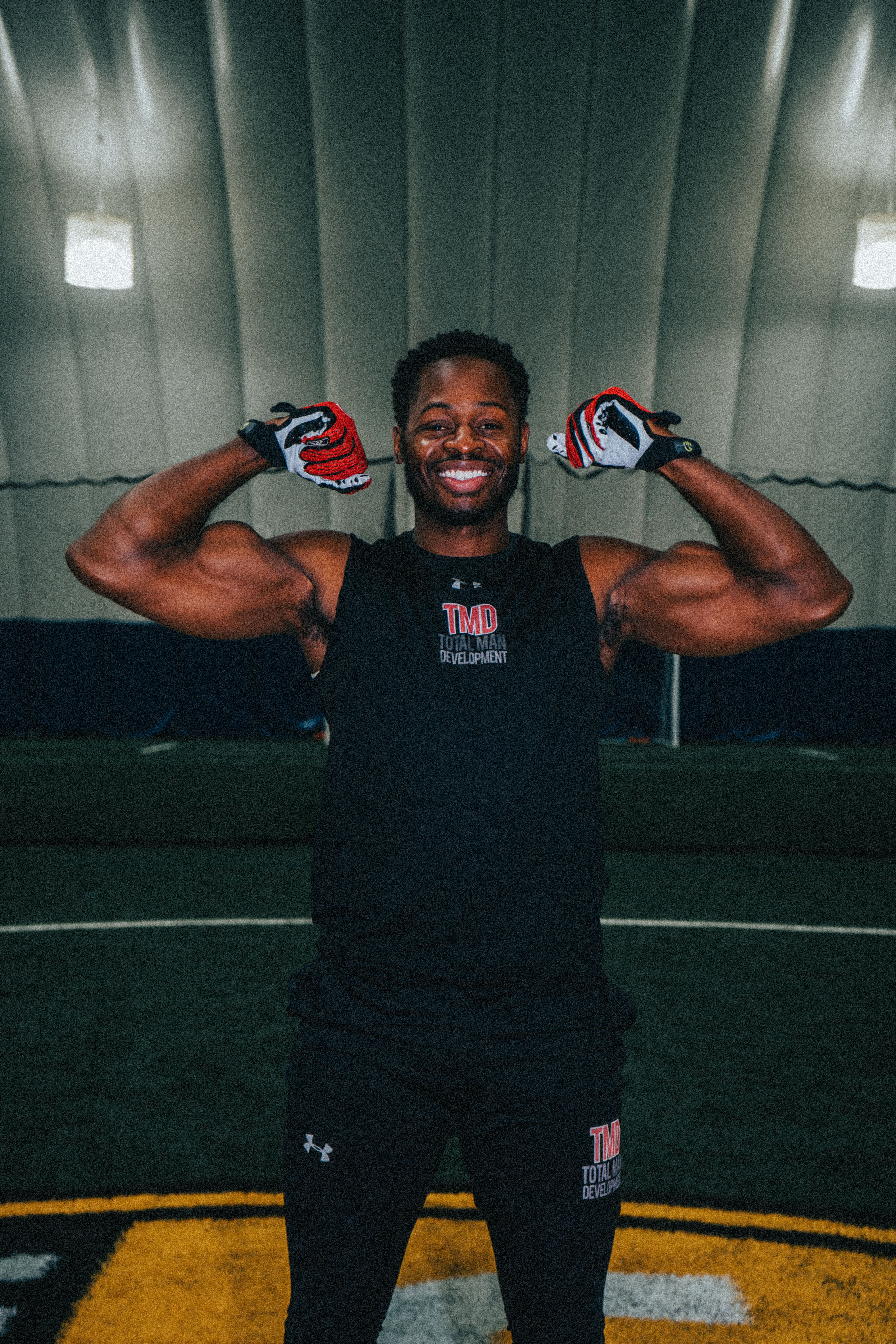 Smiling athletic man flexing muscles wearing gloves indoors, showcasing strength and fitness.
