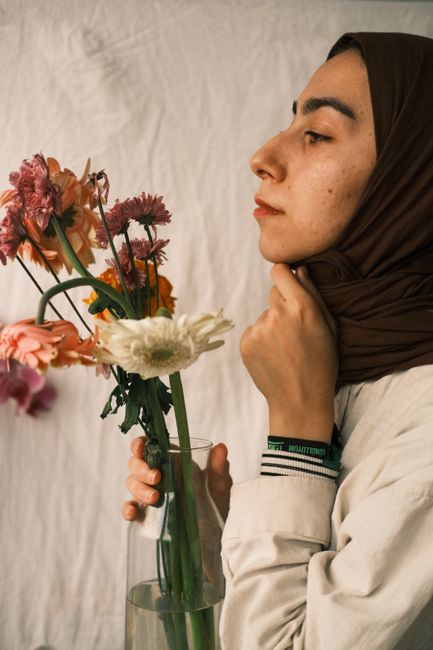 Side profile of a woman in hijab holding a vase with vibrant flowers, against a soft white background.