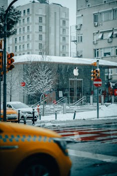 Snow-covered urban street with a modern building facade, featuring taxis and traffic lights in winter.