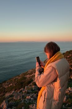 A woman in warm clothing photographs a stunning ocean sunset using her smartphone.