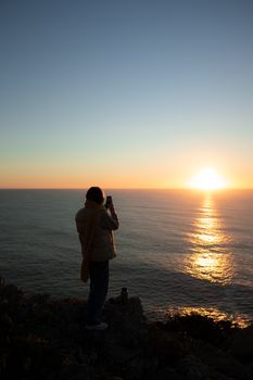 A person silhouetted against a beautiful ocean sunset, capturing the scene with a smartphone.
