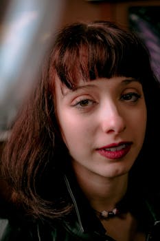 Close-up portrait of a young woman with dark hair and lipstick, indoors with soft lighting.
