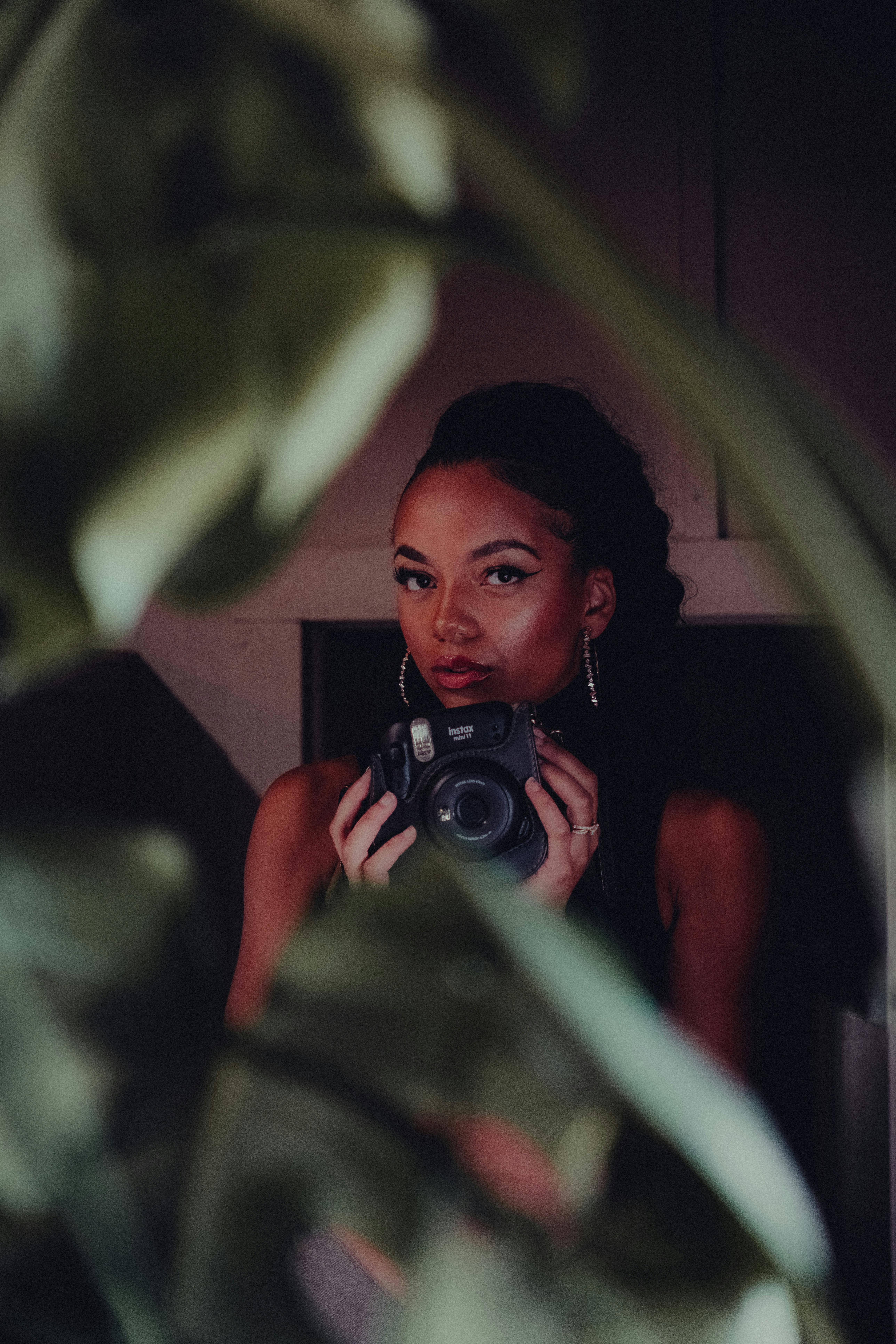 Artistic portrait of a black woman holding a camera behind blurred leaves, adding a mysterious touch.