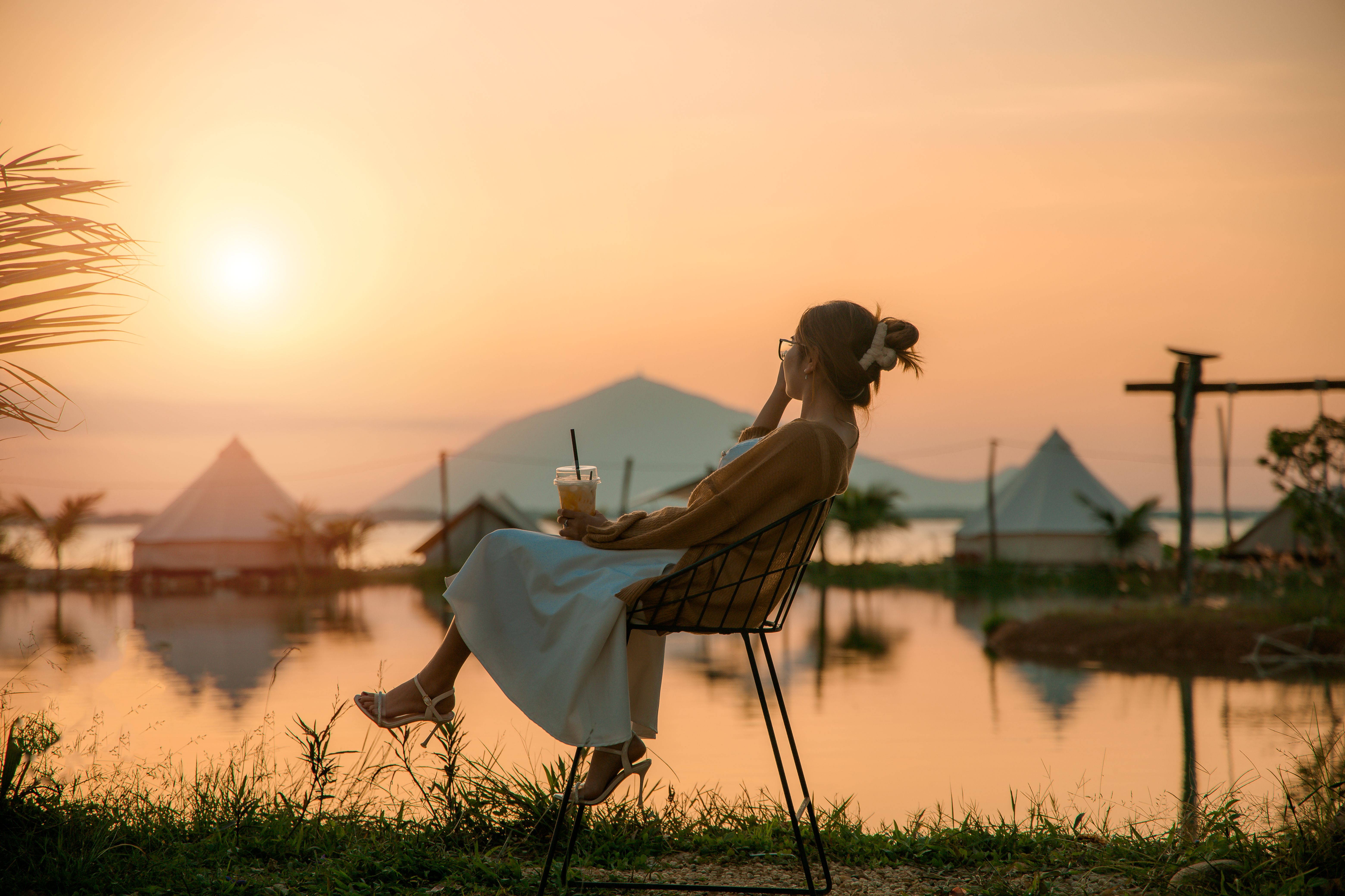 A woman enjoys a peaceful sunset by the lake, sipping a beverage.