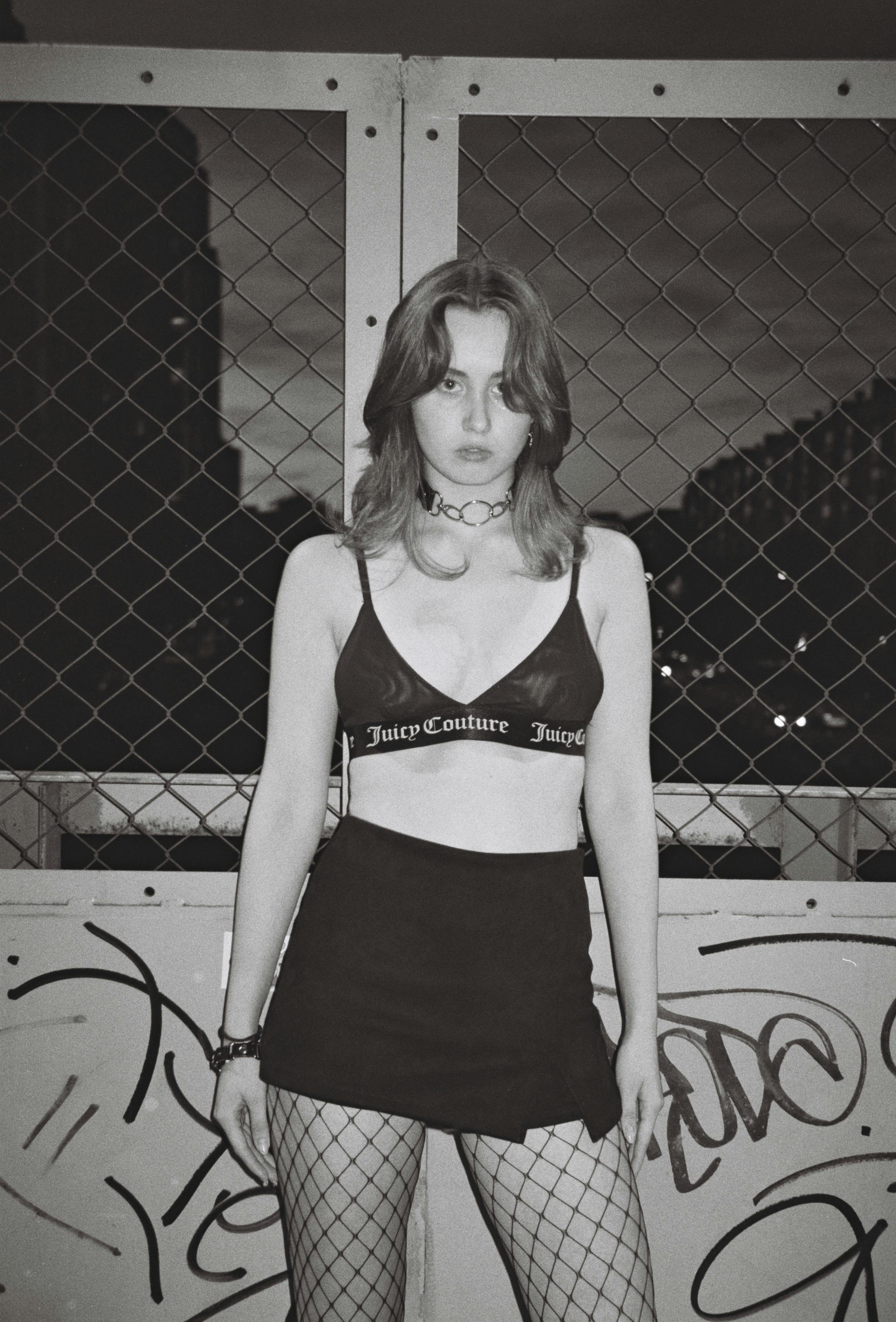 grayscale photography of a beautiful woman standing near metal chain link fence while seriously looking at the camera