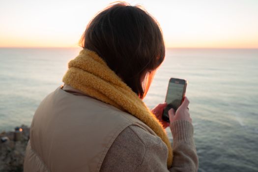 Woman using a smartphone at sunset by the sea, feeling serene and connected.