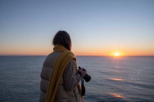 A woman photographs the serene sea sunset, capturing the vibrant horizon.