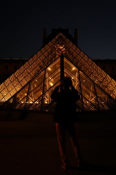 A silhouette of a person capturing the illuminated Louvre Pyramid at night in Paris.
