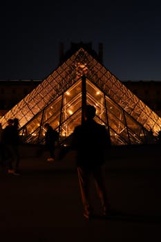A night view of the Louvre Pyramid in Paris, captured in warm light with silhouettes.