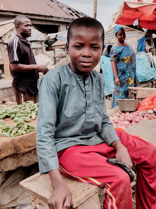 Free A young boy sits at an outdoor market, surrounded by fresh produce and vibrant activity. Stock Photo