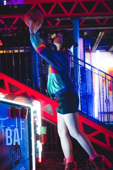 A woman enjoys a basketball arcade game under vibrant neon lights, creating a lively nightlife atmosphere.