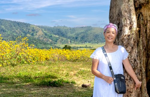Free Smiling woman in white dress with headscarf leaning on a tree in a scenic countryside landscape. Stock Photo