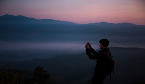 A Man in Black Jacket Standing on Mountain while Taking Picture