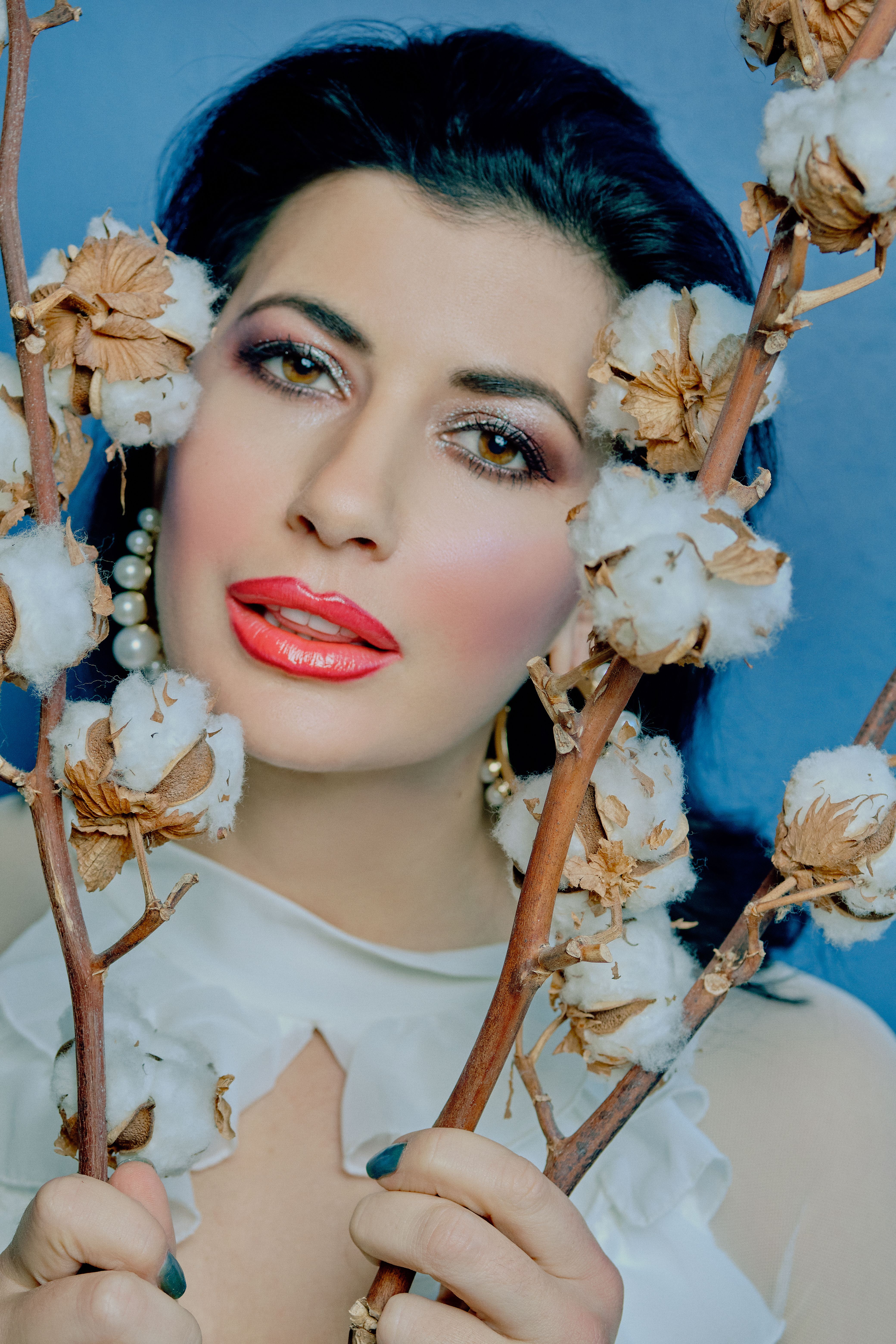 Close-up portrait of a woman with vibrant makeup framed by cotton flowers against a blue background.
