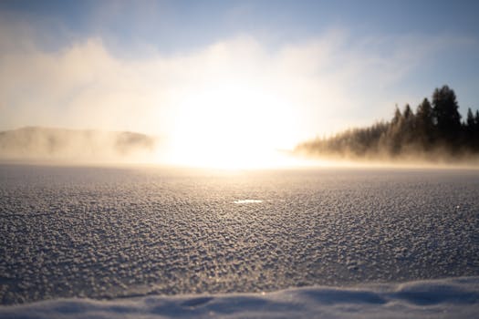 A serene winter landscape featuring a frozen lake covered in snow with mist rising during sunrise.