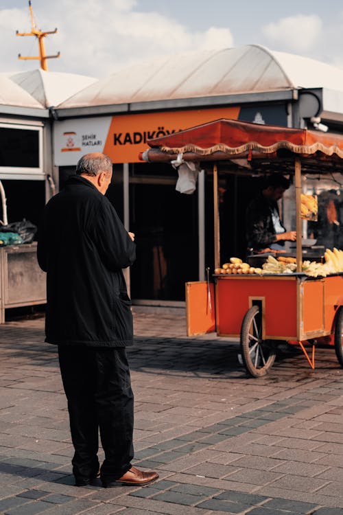 A Man in Black Jacket and Pants Standing Near the Food Cart