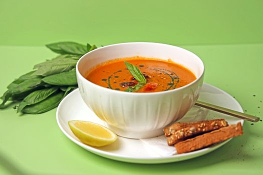 A vibrant tomato soup garnished with basil, accompanied by lemon and breadsticks.