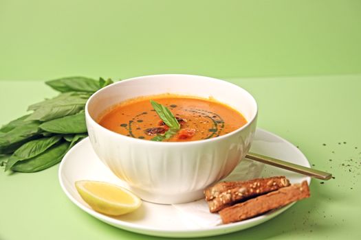 Delicious creamy tomato soup garnished with basil leaves and served with breadsticks on a green background.