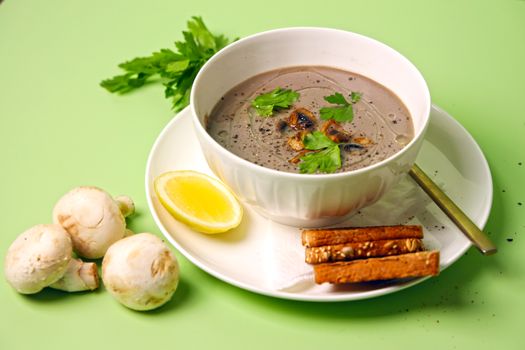 A close-up of mushroom soup garnished with parsley, served with lemon and toast on a light green background.