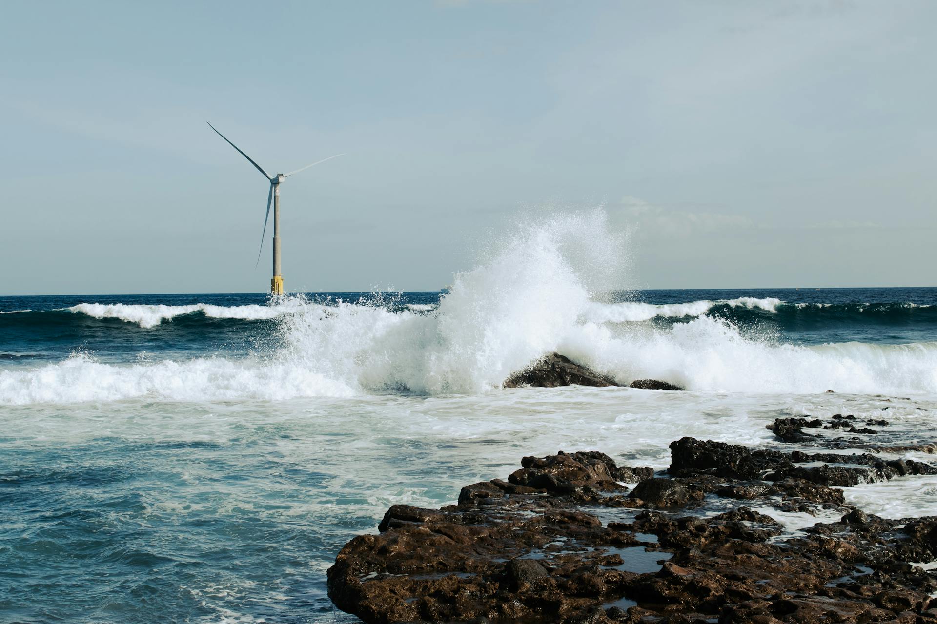 Waves crash against rocks near an offshore wind turbine on a sunny day.