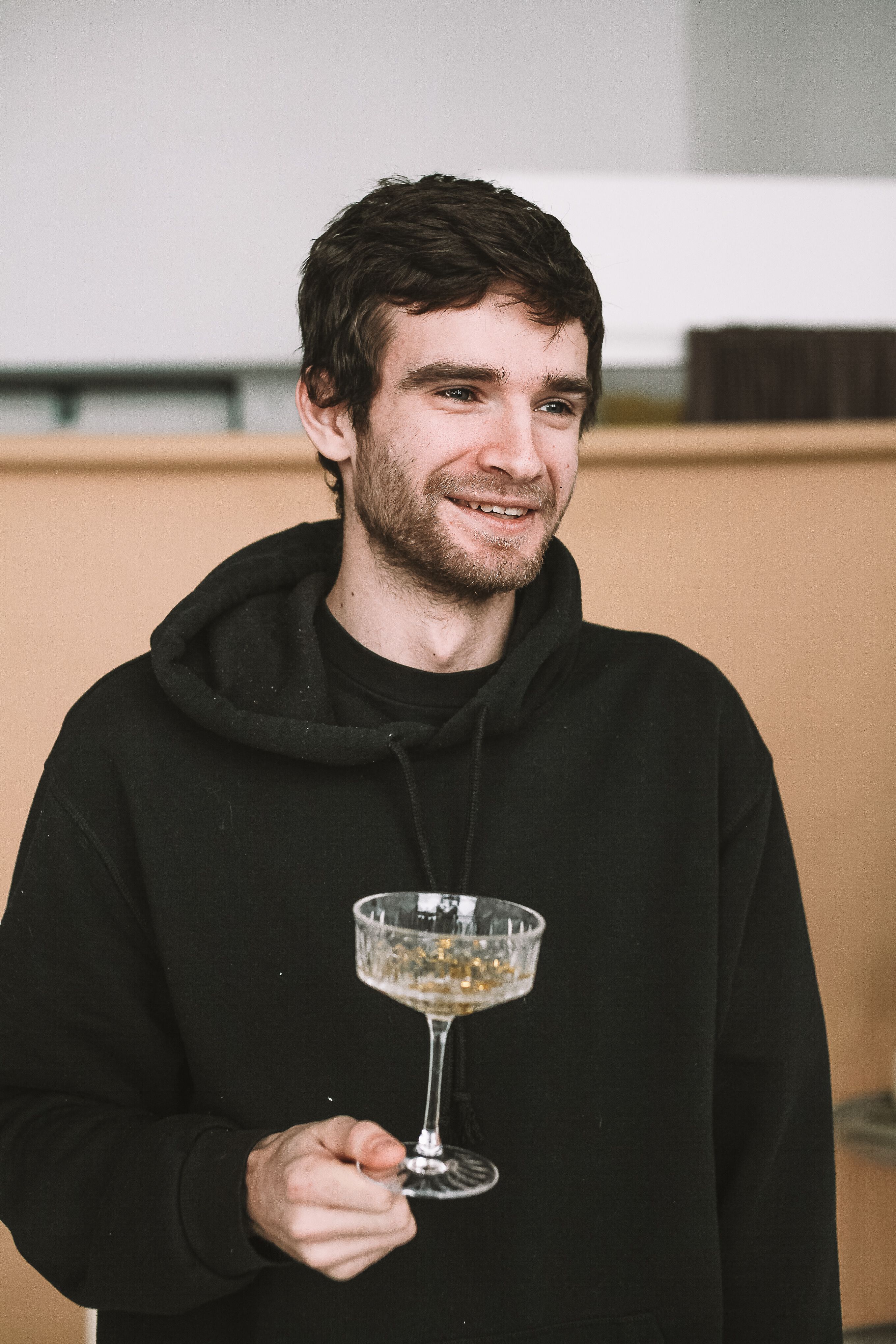 Smiling man in a black hoodie holding a champagne glass indoors.