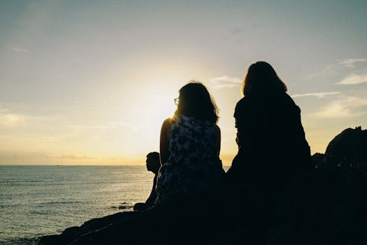 Three friends in silhouette sit on rocks, enjoying a serene sunset over the ocean.