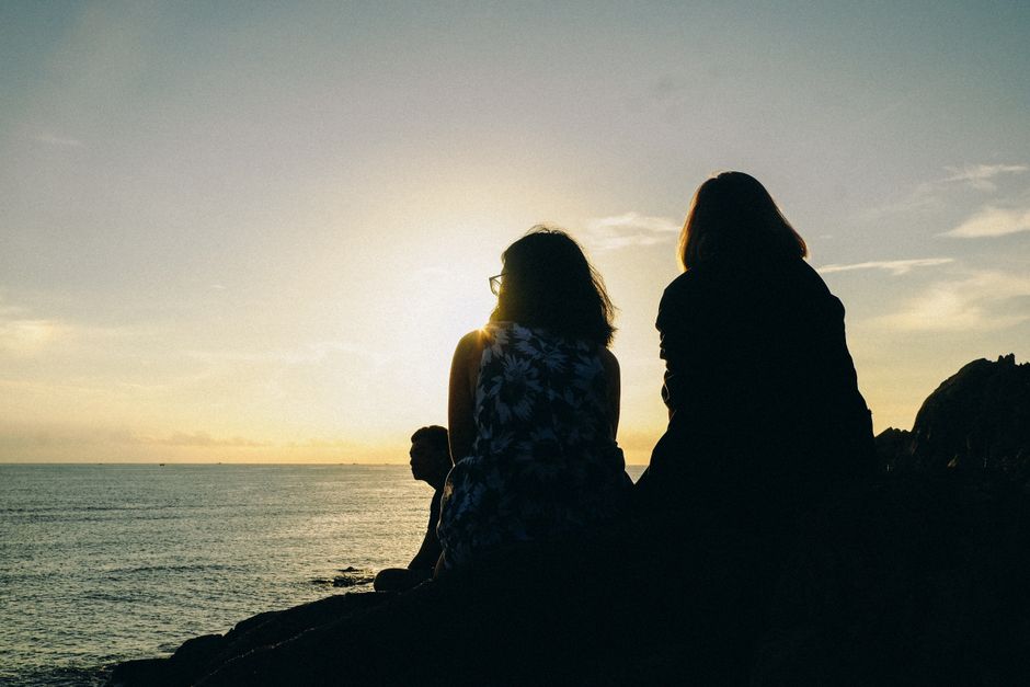 Three friends in silhouette sit on rocks, enjoying a serene sunset over the ocean.