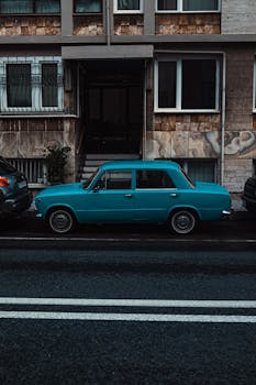 A classic blue car parked on a city street in front of residential buildings.