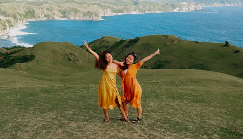 Young Women in Orange Dresses Standing on a Hill near the Shore and Smiling 