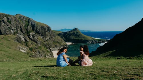 Women in Sundresses Sitting on Grass on Hill over Bay on Sea Coast