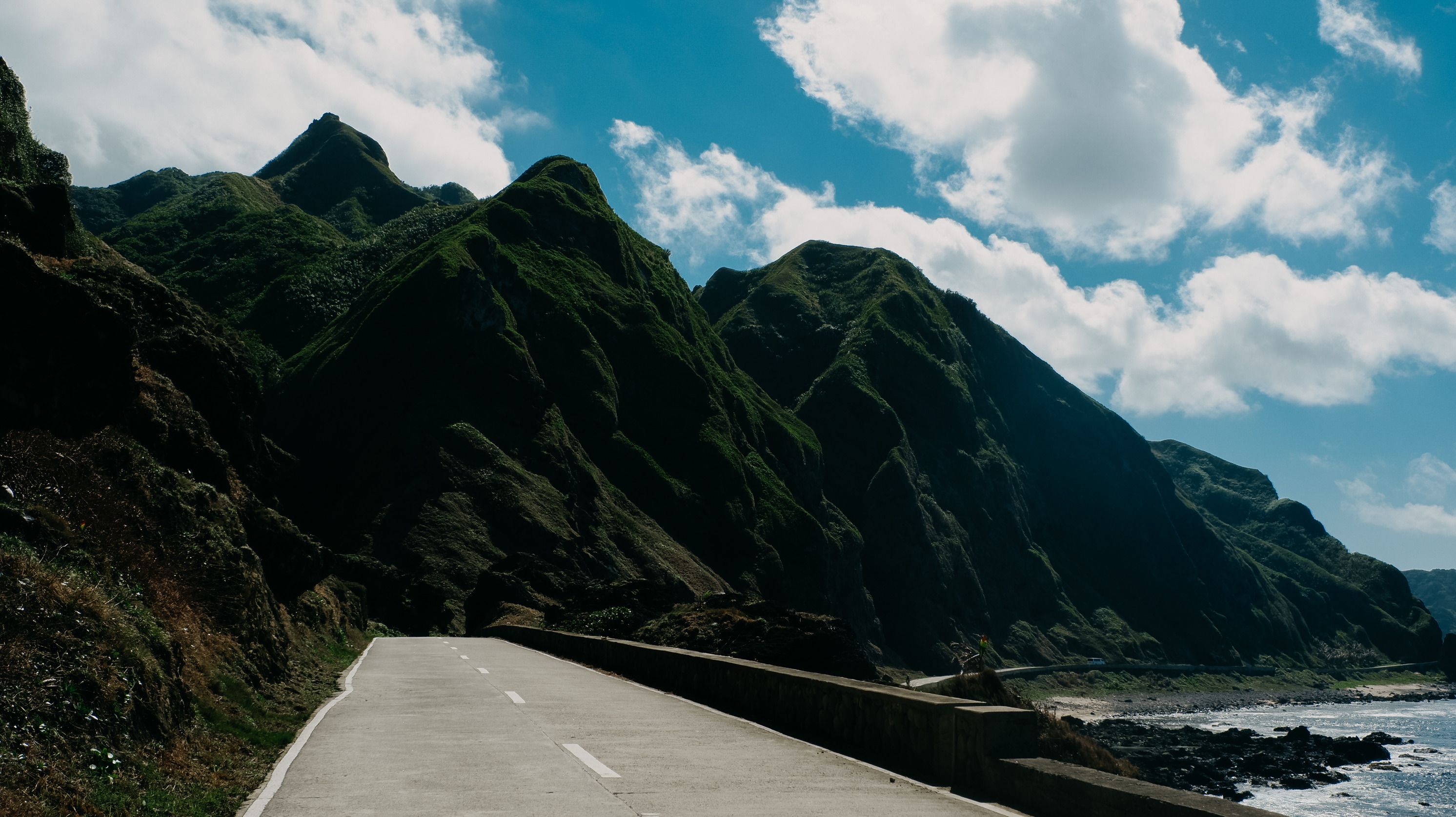 Beautiful road along lush green mountains by the coast under a vibrant blue sky.