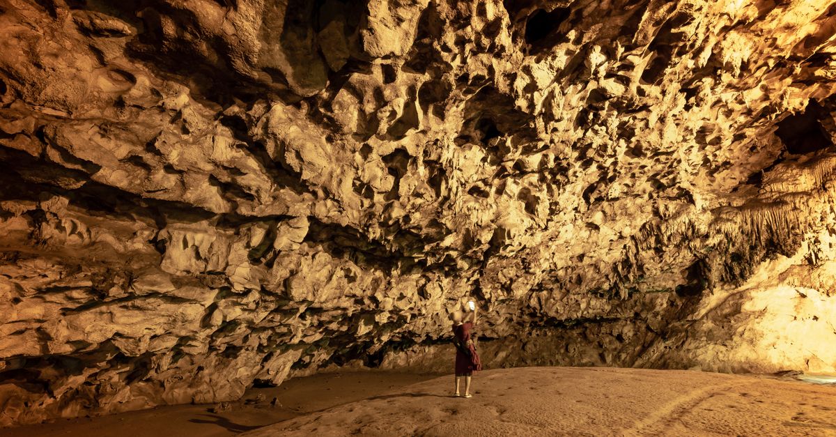 Person exploring large cave with limestone formations, illuminated by warm light.