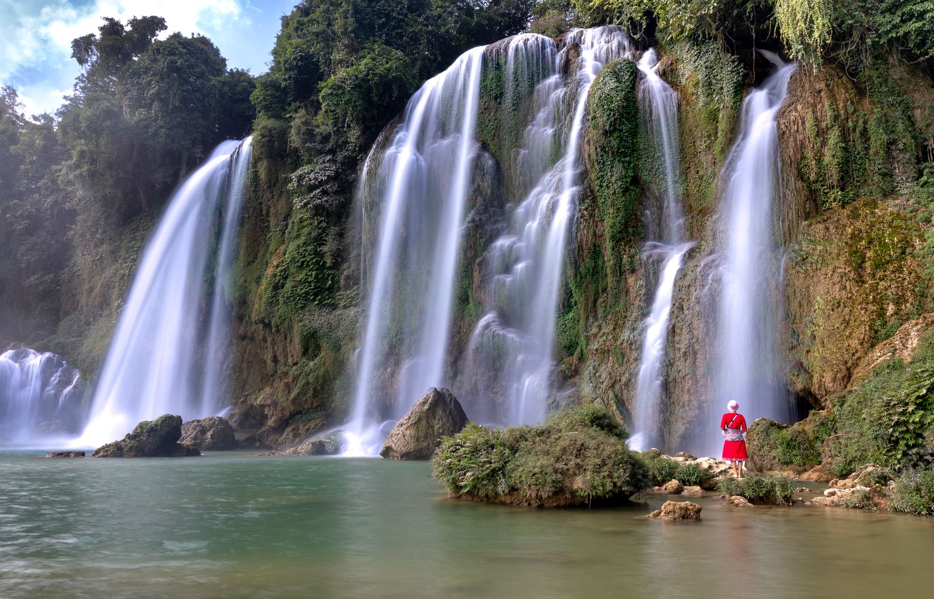 A person in red observes a stunning cascading waterfall surrounded by lush greenery.