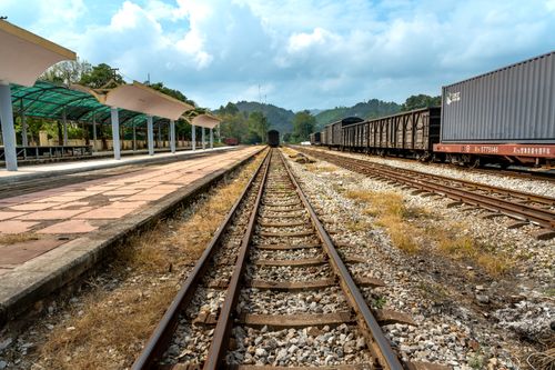 Free A cargo train waits on railway tracks at an empty outdoor station, surrounded by greenery and cloudy skies. Stock Photo