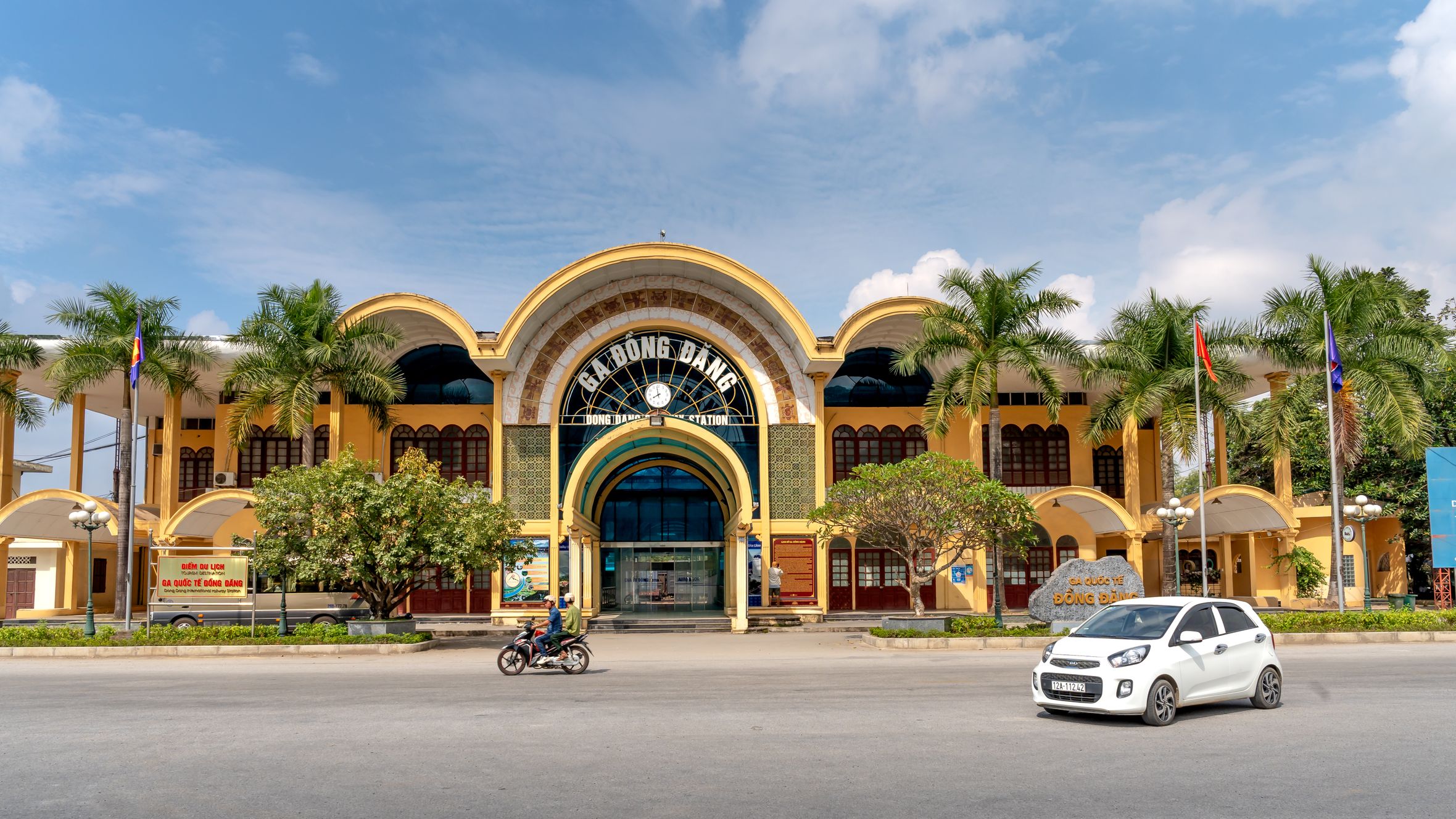 Scenic view of Dong Dang Station with palm trees, car, motorcycle, and clear sky.