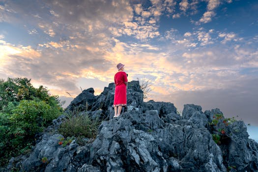A woman in a red dress stands on a rocky cliff during sunrise, with beautiful clouds overhead.