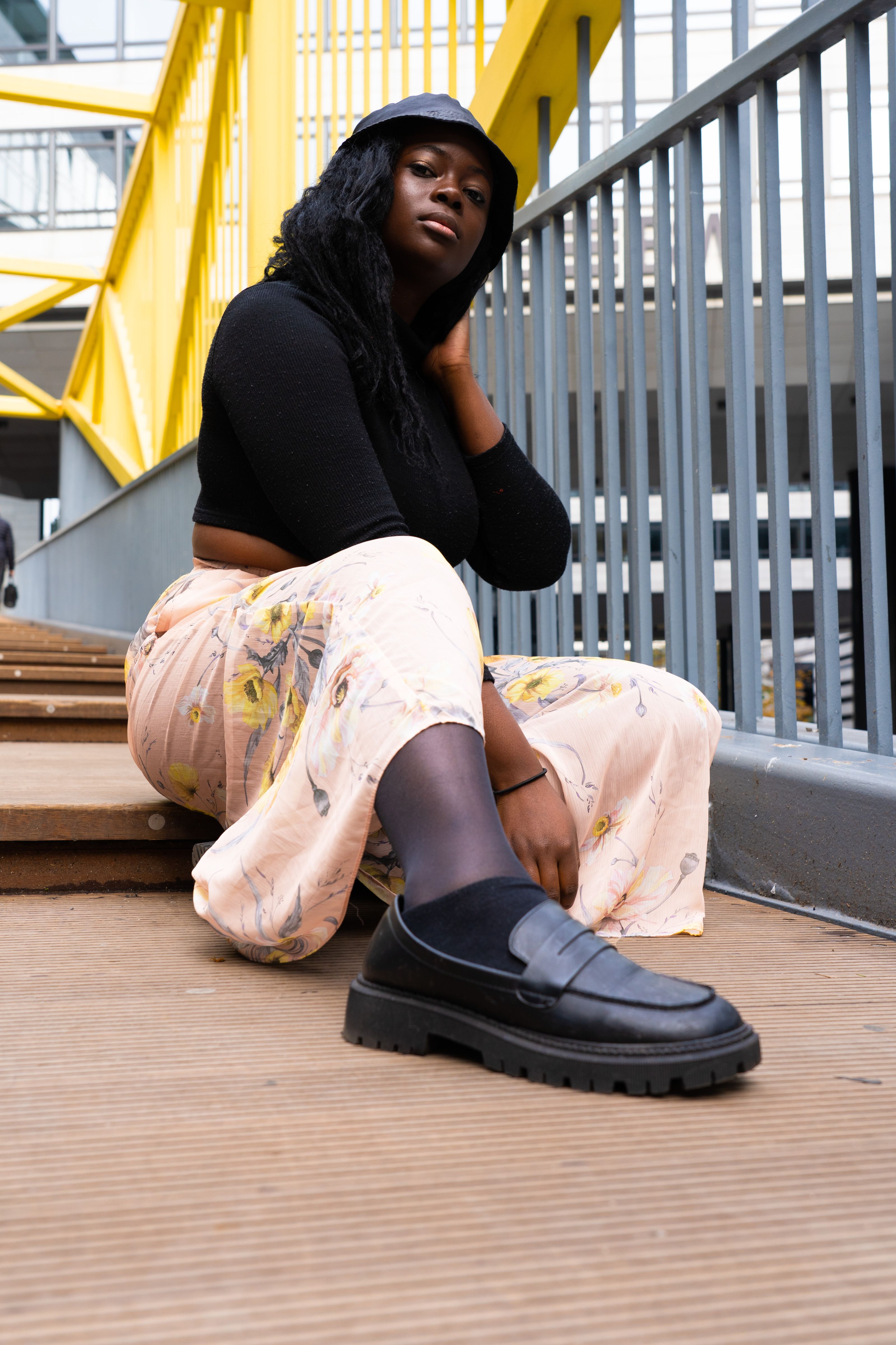 Mulher negra sentada em escadas urbanas, usando uma moda estilosa com saia floral e mocassins pretos.