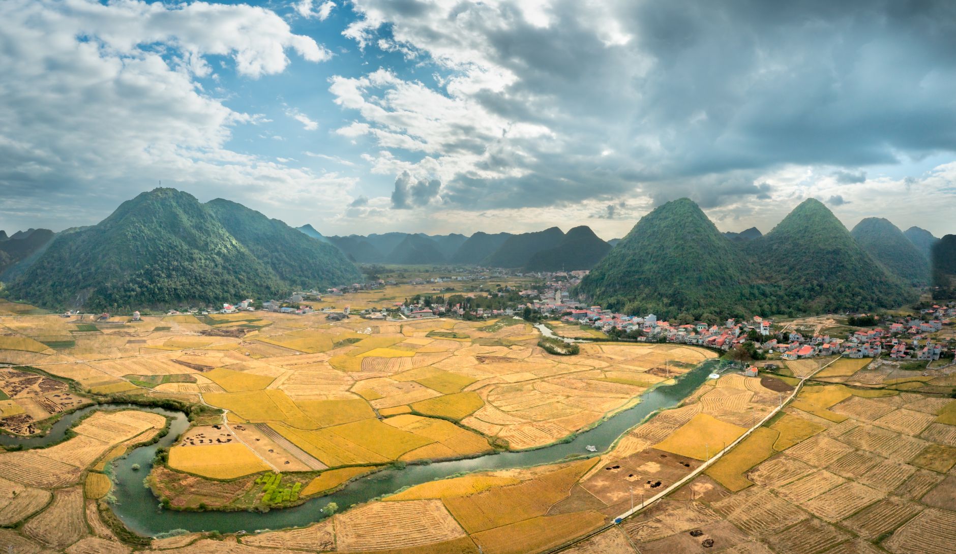 A picturesque rural landscape with rolling fields, mountains, and a river under a cloudy sky.