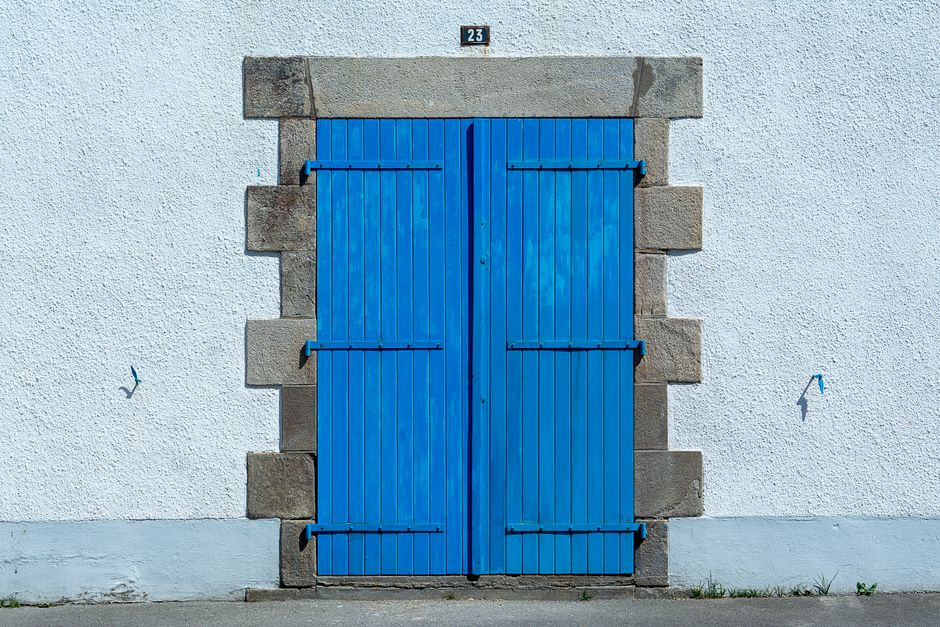 A vintage blue wooden door set in a textured white plaster wall, featuring a stone frame and house number 23.