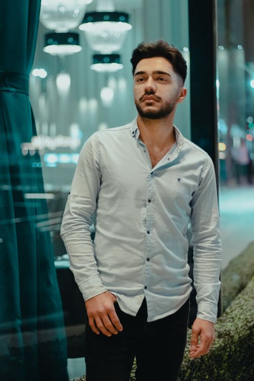 Free Portrait of a man in a button-down shirt standing in a city at night, looking thoughtful. Stock Photo
