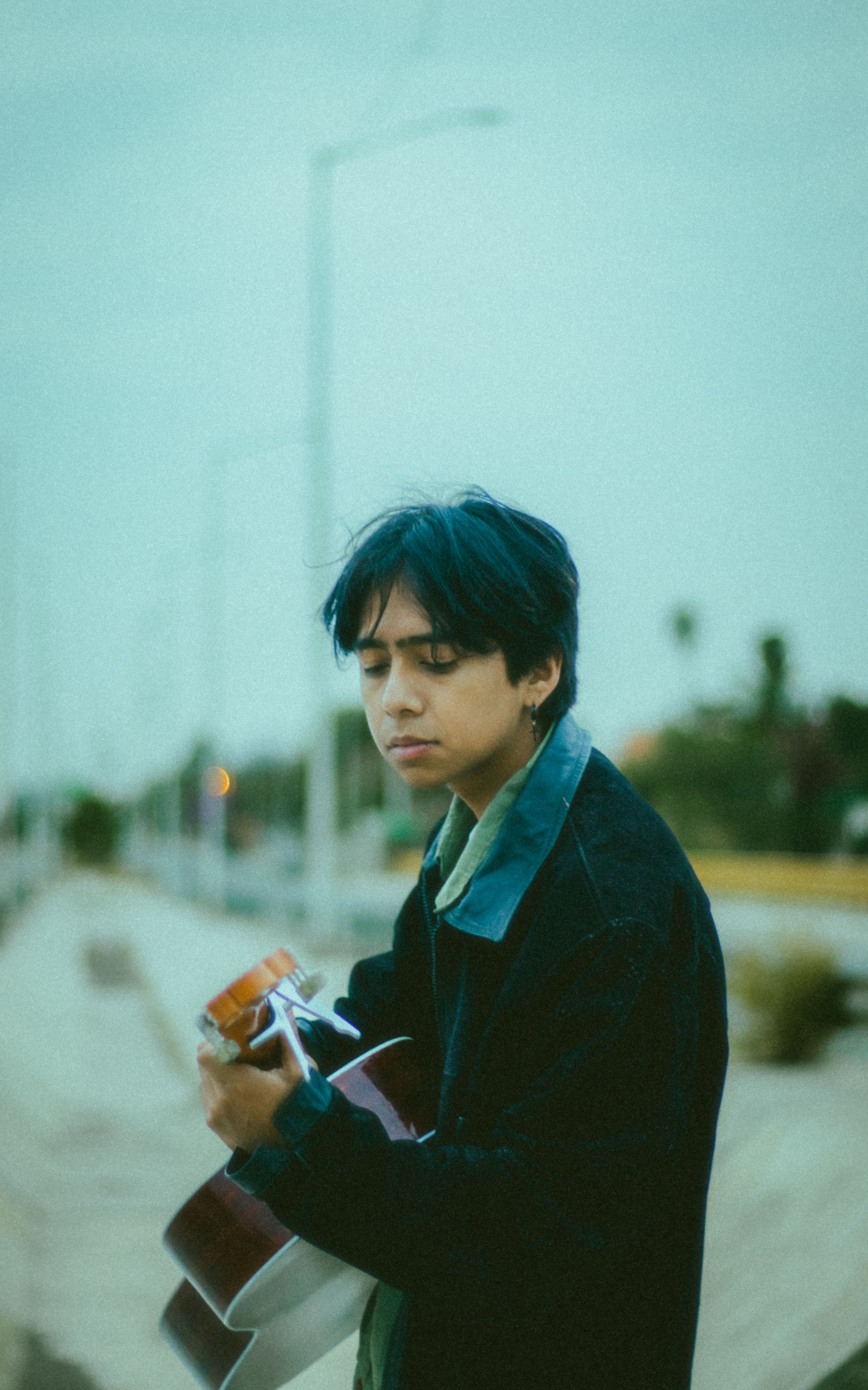 A young man in a black jacket playing guitar by the roadside on a cloudy day.