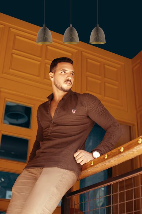 Free A fashionable man with facial hair in brown attire leaning on a wooden railing indoors. Stock Photo