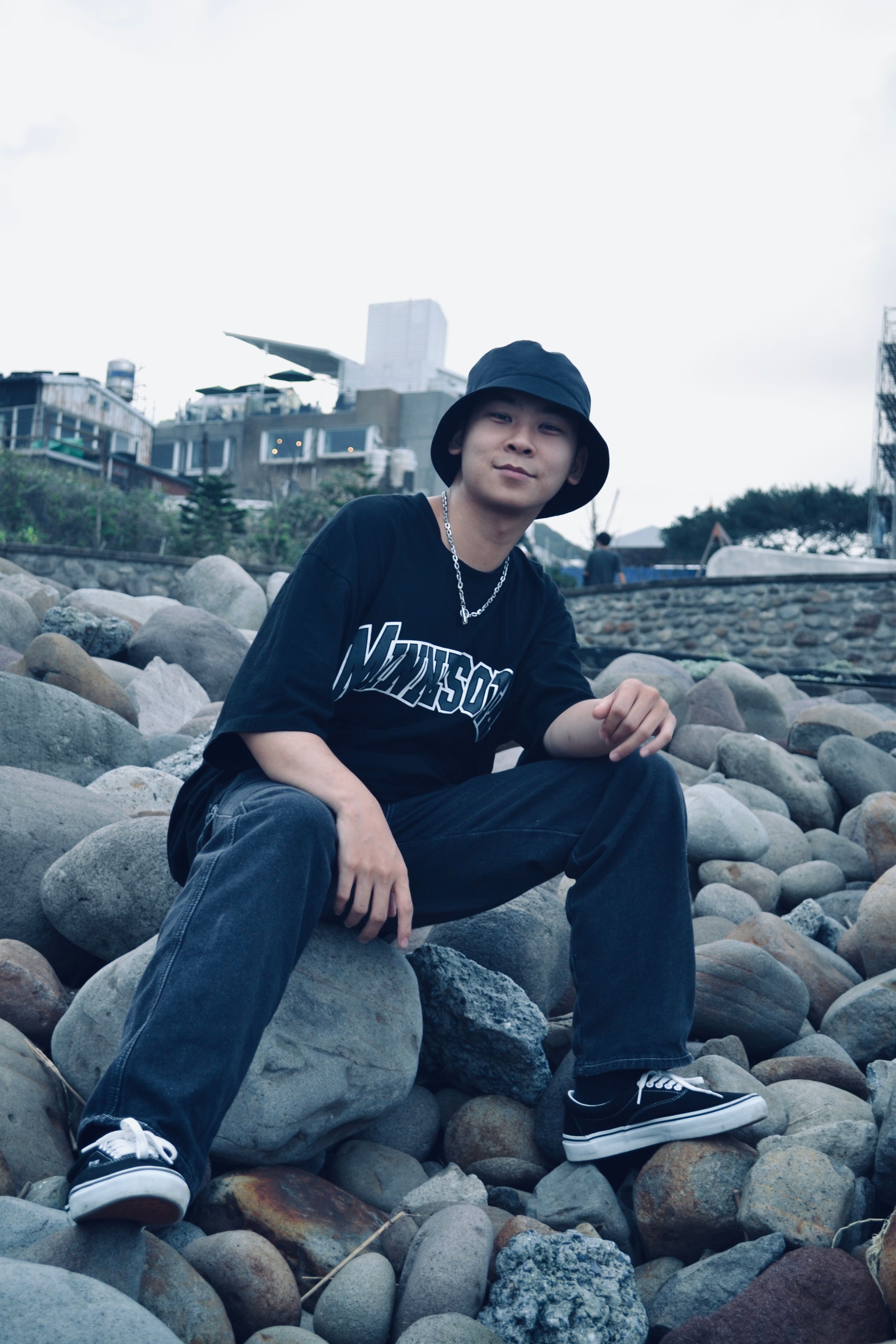 Young man sitting on rocky beach wearing a bucket hat and casual attire.