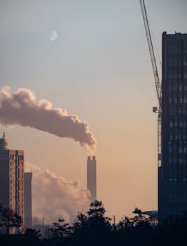 A cityscape featuring smokestacks emitting smoke, captured at sunrise with a visible moon.
