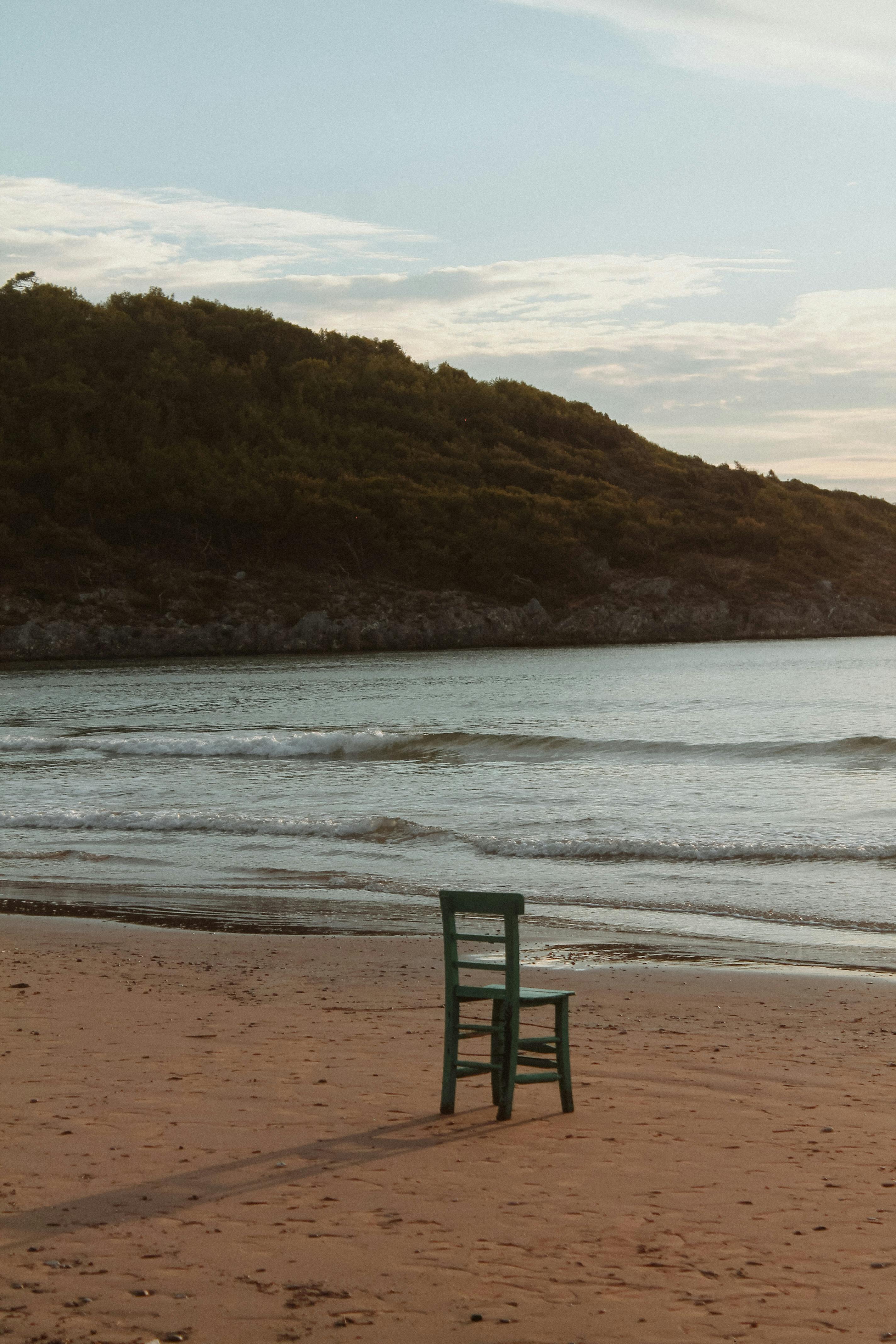 A lone chair on a tranquil beach with gentle waves and a green hillside in the background.