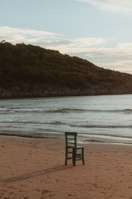 A lone chair on a tranquil beach with gentle waves and a green hillside in the background.