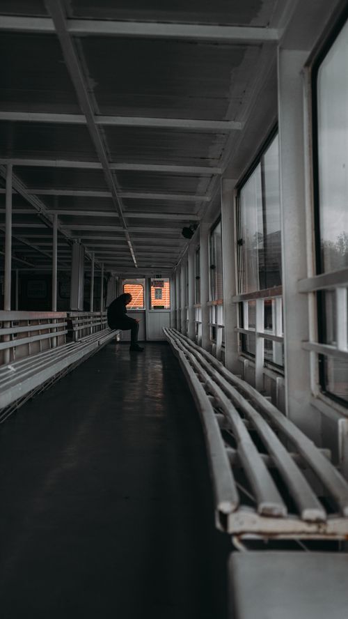Free Moody silhouette of a person sitting alone in a dimly lit empty hallway with windows. Stock Photo