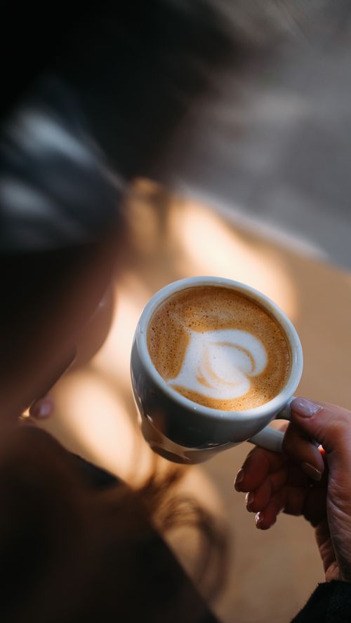 Free Close-up of a hand holding a warm cup of coffee with beautiful latte art in natural sunlight. Stock Photo